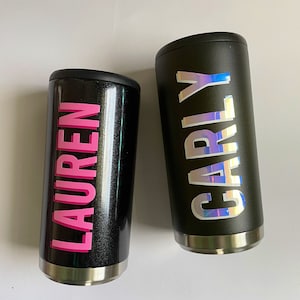 Puede incluir: Dos vasos de acero inoxidable negro con nombres personalizados. El vaso de la izquierda tiene el nombre "LAUREN" en letras rosas. El vaso de la derecha tiene el nombre "CARLY" en letras iridiscentes.