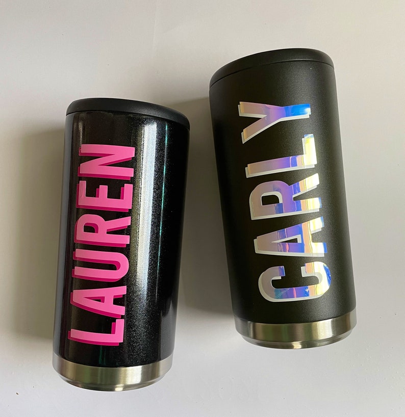Puede incluir: Dos vasos de acero inoxidable negro con nombres personalizados. El vaso de la izquierda tiene el nombre "LAUREN" en letras rosas. El vaso de la derecha tiene el nombre "CARLY" en letras iridiscentes.