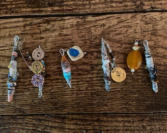 May include: Five colorful, handmade, decorative charms made of fabric, beads, buttons, and metal. The charms are arranged in a row on a wooden surface.
