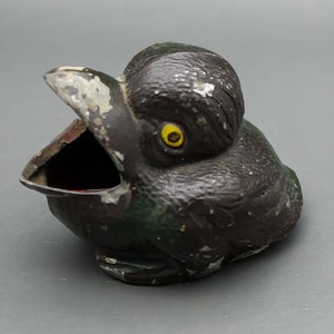May include: A vintage, black bird-shaped ashtray with an open beak. The ashtray has yellow eyes and a green underside. The paint is chipped, giving it a rustic appearance. The interior of the beak is red.