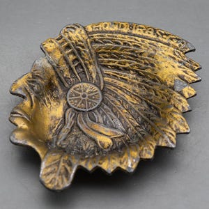 May include: A metallic gold and dark bronze-toned dish shaped like the profile of a person wearing a traditional feathered headdress. The word HONDURAS is embossed along the upper edge of the headdress, featuring intricate textures and details.