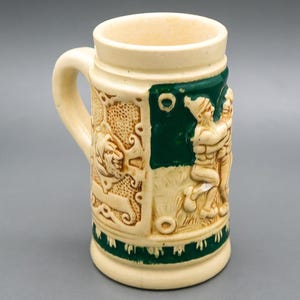 May include: Cream-colored ceramic beer stein with a handle. The stein features relief carvings of figures and a green panel. The base and top rim are also green. The stein is approximately 5 inches tall.