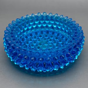 May include: A round, vibrant blue glass bowl featuring a textured hobnail pattern across its entire exterior surface. The bowl has a raised, scalloped rim and a deep, circular basin, showcasing a translucent quality that catches the light.