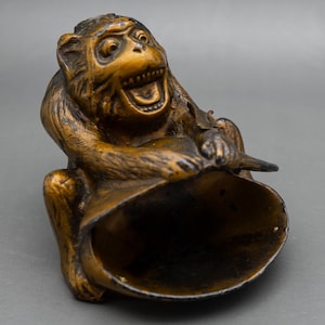 May include: A brown, carved monkey figurine with a textured, fur-like surface. The monkey is depicted in a seated position with its mouth open, holding a large, hollow, conical object that extends forward from its lap, creating a wide opening.