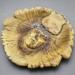 May include: A detailed, antique-style ashtray with a central face design surrounded by a feather-like pattern. The ashtray is primarily tan with darker accents, and a small shield-shaped element is visible.