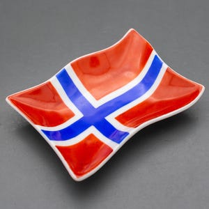 May include: A small, rectangular ceramic dish with wavy, upturned edges. The surface features a glossy finish painted with the Norwegian flag design, consisting of a bright red background, a white cross, and a bold blue cross centered on top.