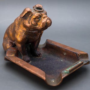 May include: A copper bulldog ashtray with a small hole on top of its head. The ashtray is rectangular with a raised edge and a small lip on one side.
