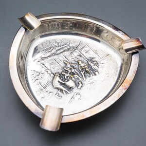 May include: A silver-colored ashtray with a triangular shape and three cigarette rests. The interior features a detailed embossed scene. The ashtray is made of metal and has a shiny finish.