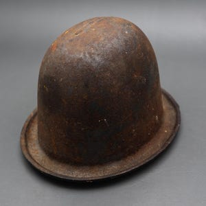 May include: A vintage, dome-shaped metal object with a wide, flat brim and a heavily rusted, dark brown, textured surface. The item features a rounded crown and a distinct, weathered appearance, showing signs of oxidation across its entire exterior.