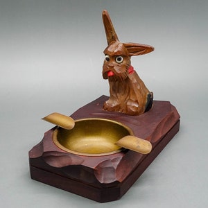 May include: Wooden ashtray with a carved dog figurine on top. The ashtray is brown and has a brass bowl. The dog is brown and has a red tongue.