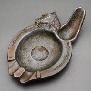 May include: A vintage ashtray with a horse head design. The ashtray is made of metal with a horseshoe shape and a small rest area. The color is a mix of bronze and dark gray.