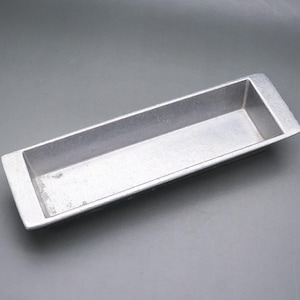 May include: A rectangular, silver-colored metal loaf pan with a slightly textured surface. The pan has a simple, utilitarian design with a raised rim on each end. The interior is smooth and the overall shape is long and narrow.