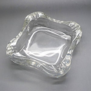 May include: A clear glass ashtray with a wavy, irregular shape. The ashtray is designed to hold cigarettes or other small items.