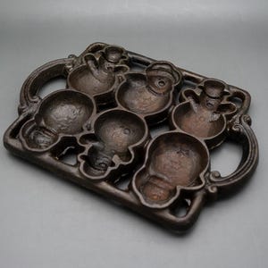 May include: A dark brown cast iron baking mold featuring six recessed cavities shaped like snowmen. The rectangular frame includes decorative scrollwork handles on both sides, with each snowman cavity displaying distinct facial and body details.