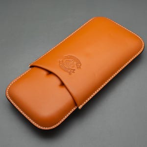 May include: A tan leather case with white stitching and a decorative emblem. The case is rectangular with rounded edges and a flap closure. The leather has a smooth texture and a warm, inviting color.