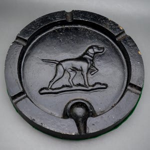 May include: Black ashtray with a raised relief of a dog. The round ashtray has a small notch for resting cigarettes and a green felt base. The dog is in a hunting pose.