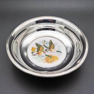 May include: A silver-colored, round bowl with a decorative center featuring a painted scene of two flying ducks. The ducks have brown, green, and white feathers, with yellow and green foliage below. The bowl has a shiny, reflective surface.