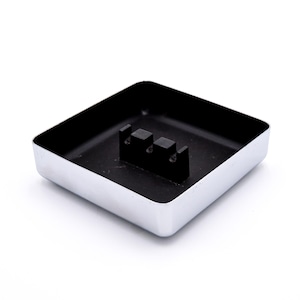 May include: Square ashtray with a shiny silver exterior and a black interior. The ashtray has a raised black section in the center with two rectangular slots. The ashtray is on a white background.