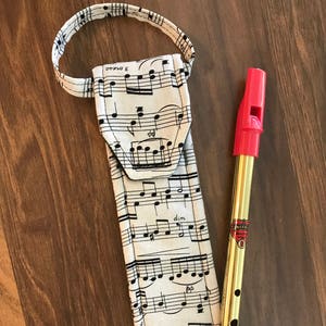 May include: A white fabric case with a musical note pattern and a red and gold tin whistle. The case has a loop handle. The whistle has a red mouthpiece and a gold body with a black label that says "Emerald, British Made".