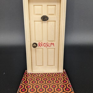 Puede incluir: Una réplica en miniatura de la icónica puerta de la película "El Resplandor". La puerta es blanca con el número "237" en negro y la palabra "REDRUM" escrita en rojo. La puerta está sobre una alfombra con estampado negro y rojo.