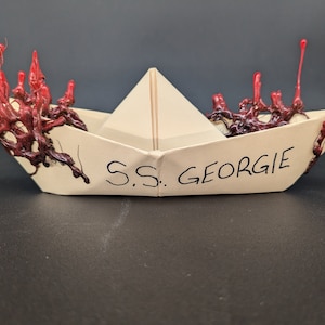 Puede incluir: Un barco de papel con cera roja y negra goteando por los lados. El barco está etiquetado como "S.S. Georgie."