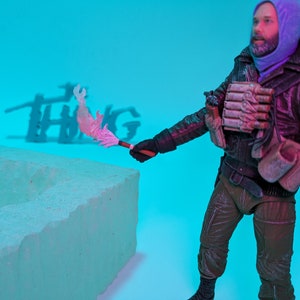 Op de afbeelding: Een figuur met een paarse capuchontrui en een bruine broek houdt een roze en witte fakkel vast voor een wit blok. De figuur staat voor een blauwe achtergrond met het woord "THING" in de schaduw achter de figuur.