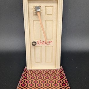 Puede incluir: Una réplica en miniatura de la puerta de la película "El Resplandor" con un hacha que sobresale. La puerta es blanca con una inscripción roja que dice "REDRUM". La puerta está sentada sobre una base con un patrón negro y rojo.