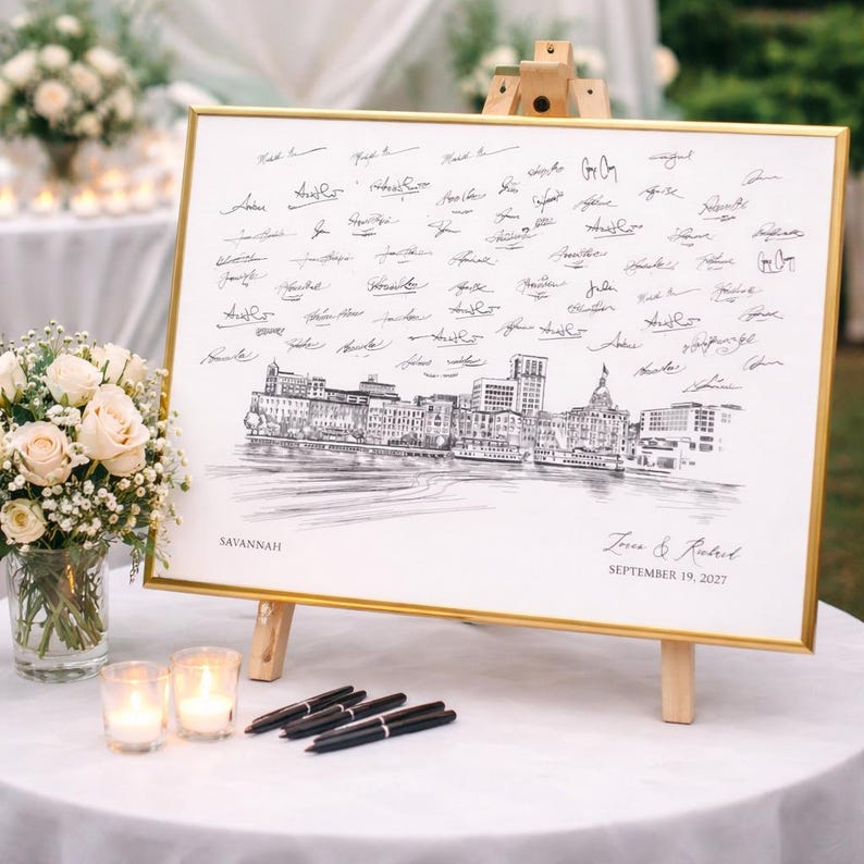 May include: A rectangular guest book sign in a gold frame featuring a black and white sketch of the Savannah skyline. The print includes the text Savannah, the names Zoe & Robert, and the date September 19, 2027, surrounded by many handwritten signatures.