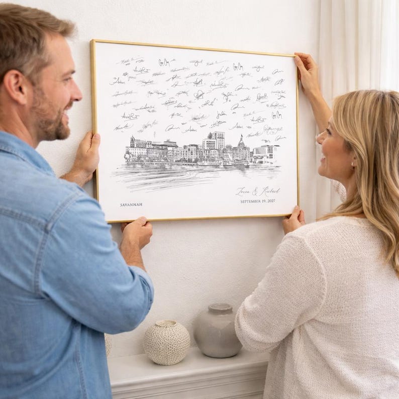 May include: A rectangular wall art print in a thin gold frame featuring a black and white sketch of the Savannah skyline. The print includes the text Savannah, the date September 19, 2027, and the names Jane & Robert, with many signatures above.