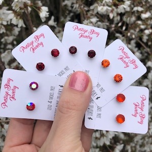 Puede incluir: Cuatro tarjetas con pares de pequeños pendientes de botón rojos, naranjas e iridiscentes. Cada tarjeta tiene el texto "Prestige Bling Jewelry" y "Lead & Nickel Free".
