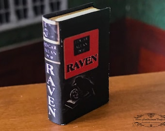 Puede incluir: Un libro en miniatura titulado "RAVEN" de Edgar Allan Poe. El libro es negro con una encuadernación roja y letras doradas. La portada presenta una silueta de un cuervo.