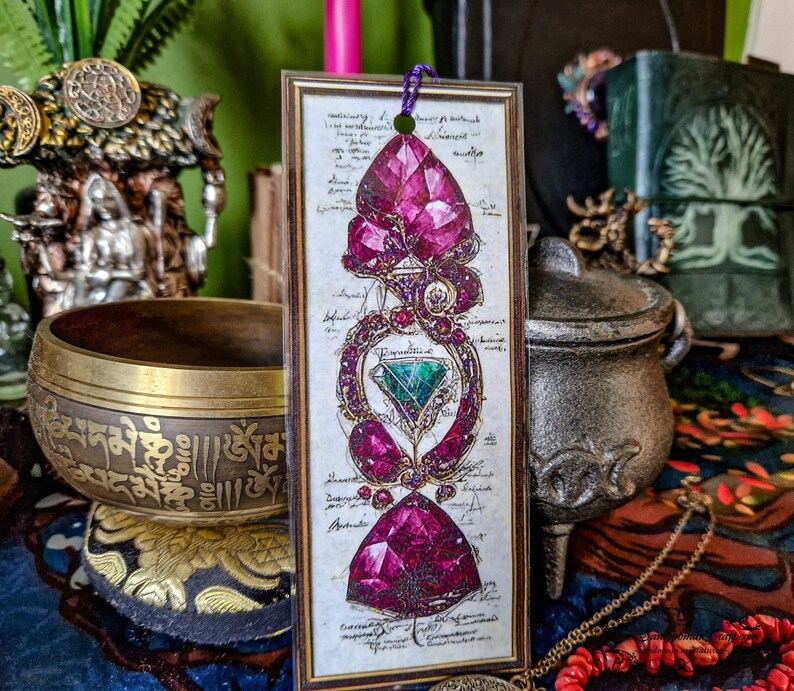 The laminated bookmark is propped up against an intricately decorated bowl and a silver pot. there are various statues and other decorative items in the background. All are sitting on a blue and red tablecloth. Large pink and turquoise stones decorate the center of the bookmark, which looks 3D against a white background. There is small, indecipherable writing on the bookmark behind the stones.