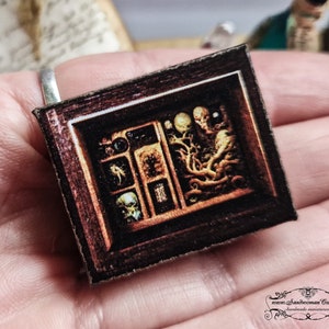 Może przedstawiać: Miniaturowe, oprawione dzieło sztuki z ciemnobrązową drewnianą ramą. Grafika przedstawia kolekcję groteskowych, nie z tego świata okazów w przegródkach. Obraz przedstawia czaszkę, dziwne stworzenia i inne niepokojące obiekty. Ogólnym tematem jest horror i makabra.