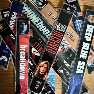 Puede incluir: Una colección de cintas VHS vintage con varios títulos de películas, como "Breakdown" y "Deep Blue Sea". Las cintas están en estuches de plástico y están dispuestas sobre una superficie de madera. Las portadas muestran ilustraciones y títulos de películas.