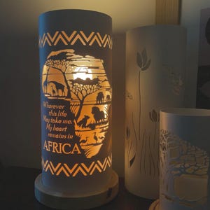 May include: A decorative cylindrical lamp featuring a cut-out African landscape with giraffes, lions, and trees. The lamp has a wooden base and a geometric pattern. Text reads "Wherever this life may take me. My heart remains in AFRICA."
