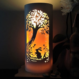 May include: A cylindrical lamp featuring a cut-out design of a tree and rabbits. The lamp emits a warm, orange glow, creating a silhouette of the tree and bunnies. The lamp is on a wooden base, with a nature and woodland theme.