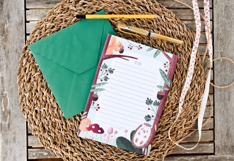 May include: A green envelope and a notepad with a woodland theme. The notepad has a white background with lined paper and a border of illustrations of a squirrel, mushrooms, berries, and a hedgehog. The notepad has the words "To Do" printed on it.