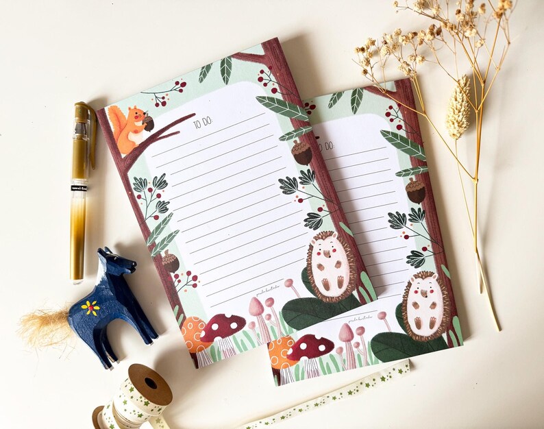 May include: Two illustrated to-do list pads with a woodland theme. The top pad features a squirrel holding an acorn and the bottom pad features a hedgehog. Both pads have a green background with brown tree branches and red berries.