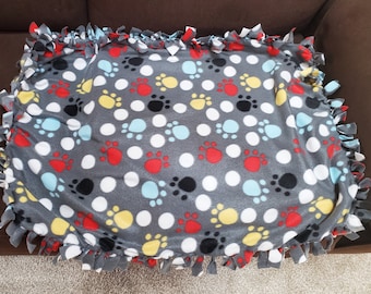 Puede incluir: Una manta de flecos de color gris de forro polar con un patrón de huellas de patas en rojo, blanco, azul, negro y amarillo. La manta tiene flecos anudados alrededor de los bordes.
