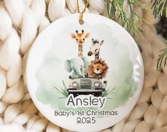 May include: A round white ceramic ornament featuring a watercolor illustration of a giraffe, zebra, elephant, and lion riding in a green safari jeep. The text reads Ansley, Baby's 1st Christmas, and 2025. It hangs from a thin gold string.