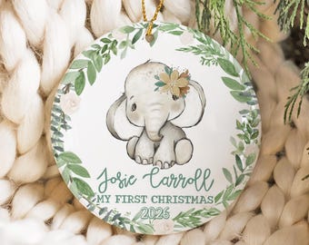 May include: A round white ceramic Christmas ornament featuring a watercolor illustration of a baby elephant with a flower on its head. The ornament is decorated with a green leaf wreath and includes the text Josie Carroll, My First Christmas, 2026.