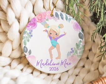 Op de afbeelding: Een rond wit keramieken ornament met een blonde gymnaste in een paars en turquoise turnpakje, omringd door een aquarel bloemenkrans van roze en paarse rozen en groene bladeren. De tekst luidt Madeline Mae en het jaartal 2026 in het paars.