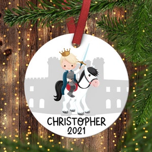 May include: White round Christmas ornament with a cartoon illustration of a prince riding a white horse with a black mane and tail. The prince is wearing a blue cape and a gold crown. The ornament has the name "CHRISTOPHER" and the year "2021" printed on it.