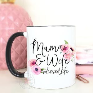 May include: White ceramic coffee mug with a black handle and black rim. The mug has a floral design and the text "Mama & Wife #blessedlife" printed on it.