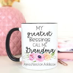 May include: White ceramic mug with a black handle and rim. The mug features the text "MY greatest Blessings CALL ME Grandma" in black script, with floral accents in pink, purple, and yellow. The name "Alina.Weston.Addison" is printed at the bottom.