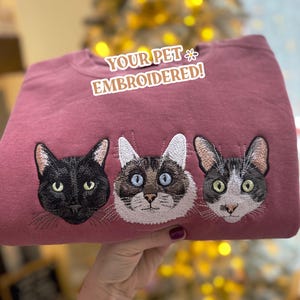 May include: A dusty rose crewneck sweatshirt featuring embroidered portraits of three cats. The text "YOUR PET EMBROIDERED!" is printed in white with a thin outline above the cat faces, which include a black cat, a Siamese mix, and a tuxedo cat.