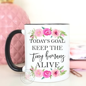 May include: White ceramic mug with a black handle and a floral design. The mug has the text "TODAY'S GOAL: KEEP THE TINY HUMANS ALIVE" printed on it.
