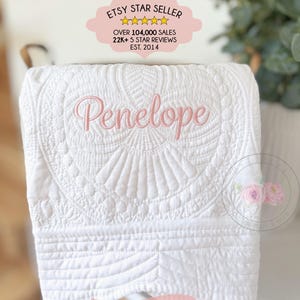 May include: A white quilted blanket featuring intricate stitched patterns and the name Penelope embroidered in soft pink script. The blanket includes a banner at the bottom with the text ADD A GIFT BOX and a top label stating ETSY STAR SELLER.