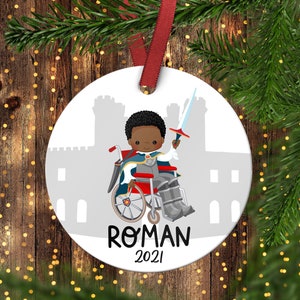 Puede incluir: Adorno de Navidad redondo blanco con una ilustración de dibujos animados de un niño negro en silla de ruedas vestido de caballero con una espada. El adorno tiene una silueta de castillo gris en el fondo y el texto "ROMAN 2021".
