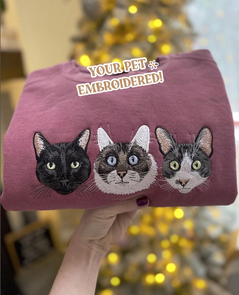 Puede incluir: Una sudadera de color malva con tres caras de gato bordadas. Los gatos son negros, marrones y blancos, y grises y blancos. El texto "YOUR PET EMBROIDERED!" est&aacute; encima de las caras de los gatos.